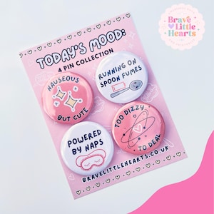 May include: A collection of four pin badges on a pink backing card, featuring the text "Today's Mood". The badges display phrases such as "Running on spoon fumes" and "Too dizzy to deal". The design includes hearts and stars.