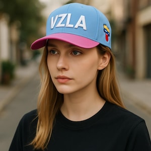 Puede incluir: Gorra de béisbol azul claro con visera rosa. La gorra muestra las letras blancas "VZLA" en la parte delantera y un pequeño emblema en el lateral. La persona lleva una camiseta negra.