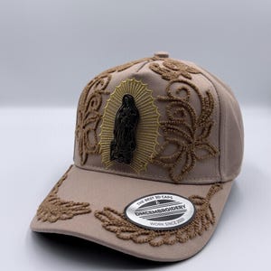 Puede incluir: Gorra de béisbol color beige con intrincados bordados marrones. La gorra presenta un emblema central de una figura religiosa y el texto "THE BEST 3D CAPS DMEMBROIDERY WORK SINCE 2020".