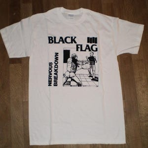 Puede incluir: Una camiseta blanca de manga corta con un estampado gráfico negro en la parte delantera. El diseño incluye el texto "BLACK FLAG" y "NERVOUS BREAKDOWN" junto a una ilustración en blanco y negro de dos figuras en una habitación con suelo ajedrezado.