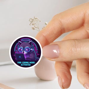 Op de afbeelding: Een ronde sticker met een wasbeer met een bril die op een laptop werkt. De wasbeer is paars met neonblauwe en roze details. De tekst "Vibe Coding Mode: Activated" staat eronder.