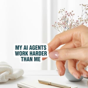 May include: A white sticker with the text "MY AI AGENTS WORK HARDER THAN ME" in dark teal. The sticker is held by a hand with light pink nail polish. A white notebook, pen, and small vase are in the background.
