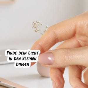 May include: A sticker with the text "FINDE DEIN LICHT IN DEN KLEINEN DINGEN" in black font. The sticker is held by a person's hand, with a small white vase and a sprig of dried flowers in the background.