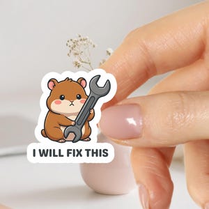 Peut inclure: Autocollant représentant un hamster de dessin animé tenant une clé. Le hamster est brun avec un ventre blanc et des joues roses. Le texte "I WILL FIX THIS" est en dessous.
