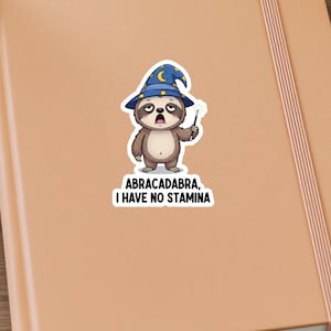 Può includere: Un adesivo con un bradipo dei cartoni animati con un cappello da mago con una luna crescente e stelle. Il bradipo tiene in mano una bacchetta e il testo recita "Abracadabra, I have no stamina."