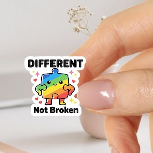 Może przedstawiać: Naklejka z tęczową postacią puzzla i napisem "DIFFERENT Not Broken". Postać ma uśmiechniętą twarz i serca. Naklejka jest trzymana przez osobę.