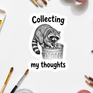 Può includere: Un adesivo con un'illustrazione in bianco e nero di un procione su un bidone della spazzatura. Il testo "Collecting my thoughts" è sopra e sotto. Circondato da materiali artistici.