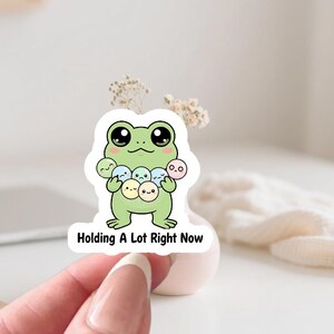 Op de afbeelding: Een sticker met een cartoonkikker in lichtgroen, met grote zwarte ogen, die verschillende kleine, ronde, pastelkleurige karakters vasthoudt. De tekst "Holding A Lot Right Now" staat eronder.
