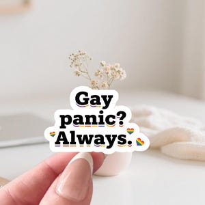 May include: A white sticker with the text "Gay panic? Always." in black, with rainbow accents. The sticker is held by a person's hand. A small vase with white flowers is in the background.