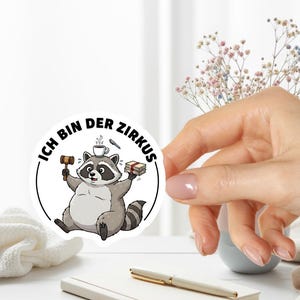 May include: A round sticker featuring a cartoon raccoon holding a gavel and books, with a coffee cup on its head. The text "Ich bin der Zirkus" is printed around the raccoon.
