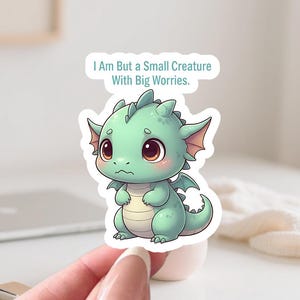 Può includere: Un adesivo raffigurante un drago dei cartoni animati in tonalità di verde con grandi occhi marroni e piccole ali. L'adesivo ha il testo "I Am But a Small Creature With Big Worries". Il drago è seduto con un'espressione preoccupata.