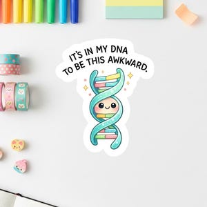 Könnte beinhalten: Ein Aufkleber mit einem Cartoon-DNA-Strang in Blau-, Rosa- und Gelbtönen mit einem lächelnden Gesicht. Der Text "IT'S IN MY DNA TO BE THIS AWKWARD" steht in schwarzer, geschwungener Schrift über dem DNA-Strang. Weitere Schreibwarenartikel sind sichtbar.