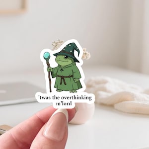 May include: A sticker of a green frog wizard with a starry hat and staff with a blue orb. The text on the sticker says, "'twas the overthinking m'lord."