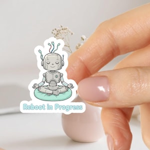 Könnte beinhalten: Ein Aufkleber mit einem Cartoon-Roboter in meditativer Pose, der auf einem hellblauen Kissen sitzt. Der Roboter ist grau mit einem silbernen Körper und hat drei Drähte, die aus seinem Kopf ragen. Der Text "Reboot in Progress" steht darunter.