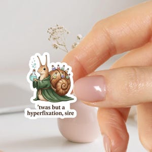 Op de afbeelding: Een sticker met een cartoon slak in een groene mantel, met een blauwe toverdrankfles. De slakkenhuis is versierd met kleine toverdrankflesjes. De tekst luidt: "'twas but a hyperfixation, sire."