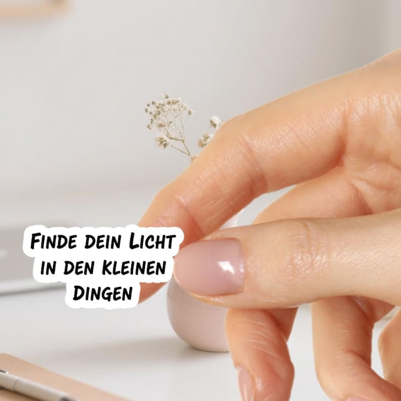 May include: A sticker with the text "FINDE DEIN LICHT IN DEN KLEINEN DINGEN" in black font. The sticker is held by a person's hand, with a small white vase and a sprig of dried flowers in the background.