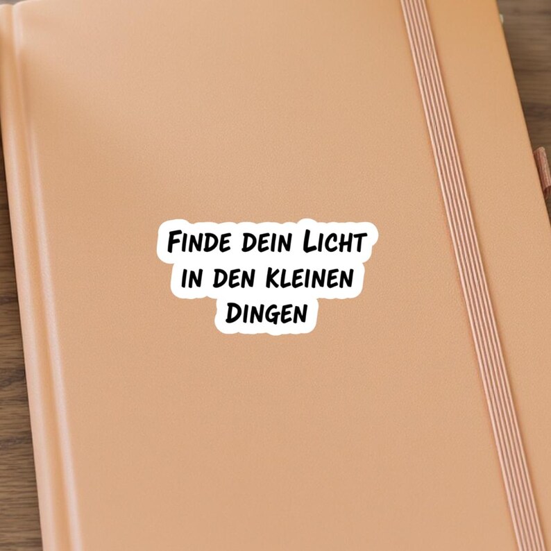 May include: A sticker with the German phrase "FINDE DEIN LICHT IN DEN KLEINEN DINGEN" in black text on a white background. The sticker is on a light brown notebook.