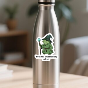 May include: A sticker showing a cartoon frog wizard in a green robe and hat, holding a staff with a blue orb. The text on the sticker says, "'twas the overthinking m'lord."