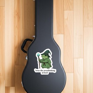 May include: A sticker depicting a cartoon frog in a wizard's outfit, including a green robe and a hat with white spots, holding a staff with a blue orb. The text says, "'twas the overthinking m'lord."