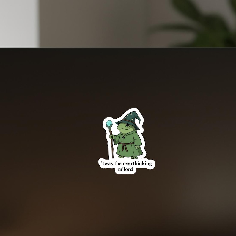 May include: A sticker depicting a cartoon frog in a wizard's outfit, including a green robe and a star-patterned hat. The frog holds a staff with a blue orb. The text on the sticker says, "'twas the overthinking m'lord."