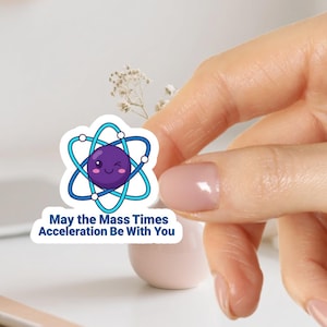 Pode incluir: Um autocolante branco com um desenho de átomo de desenho animado, com um rosto a piscar e anéis orbitais azuis e azul claro. O texto no autocolante diz "May the Mass Times Acceleration Be With You".