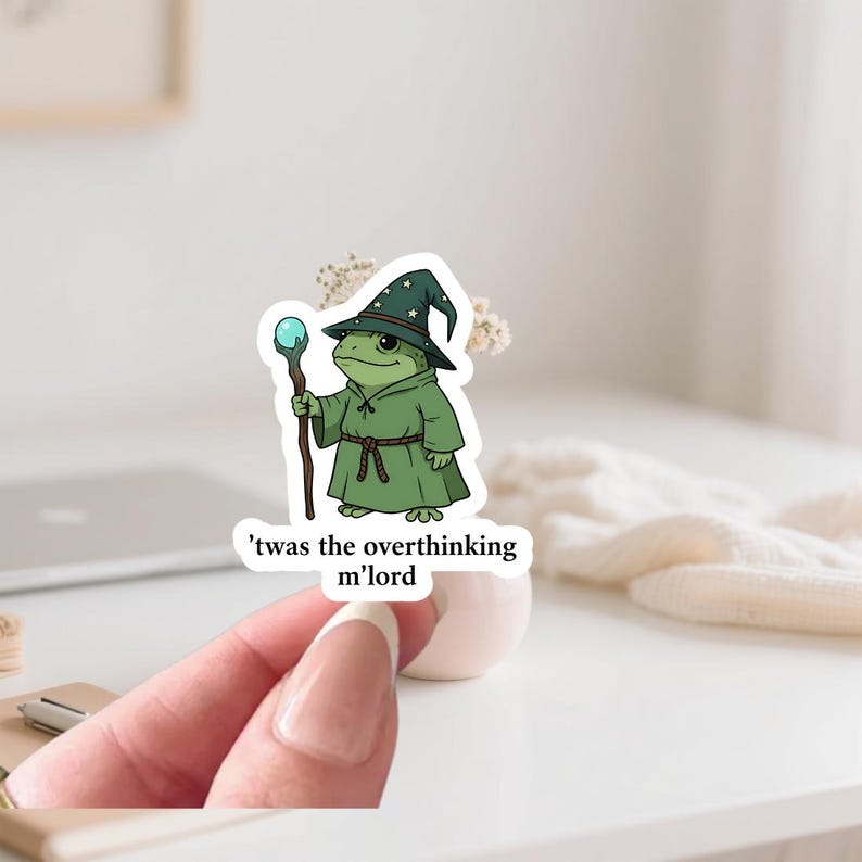 May include: A sticker of a green frog wizard with a starry hat and staff with a blue orb. The text on the sticker says, "'twas the overthinking m'lord."