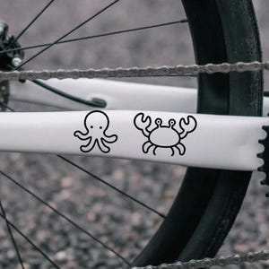 Peut inclure: Un tube de cadre de vélo blanc orné de deux autocollants en ligne noire. Le premier représente une pieuvre simple avec huit tentacules et deux yeux, tandis que le second montre un crabe avec deux grosses pinces, six pattes et deux yeux.