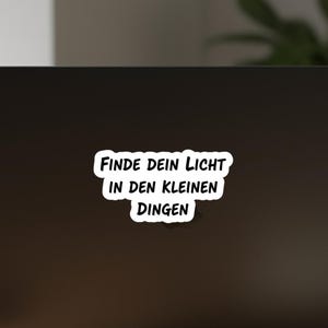 May include: A white sticker with black text that reads "FINDE DEIN LICHT IN DEN KLEINEN DINGEN". The sticker has a black border and is placed on a dark surface.