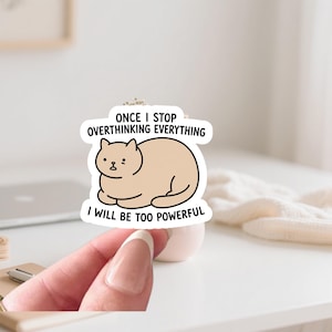 Może przedstawiać: Naklejka z kreskówkowym kotem w kolorze jasnobrązowym. Kot siedzi z tekstem "ONCE I STOP OVERTHINKING EVERYTHING I WILL BE TOO POWERFUL" w białym dymku.