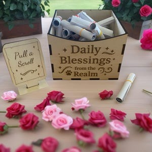 May include: A wooden box with the text "Daily Blessings from the Realm" holds rolled scrolls. A sign reads "Pull a Scroll." Red and pink roses are scattered on the table. Green plants and more roses are in the background.