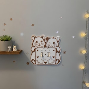 May include: A white and brown double light switch cover featuring two cartoon hamsters holding the plate. The design includes small paw prints, stars, and heart motifs etched onto the surface. The switch plate is mounted on a wall.