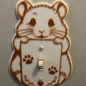 Könnte beinhalten: Eine weiße und kupferfarbene Lichtschalterabdeckung aus Kunststoff in Form eines Hamsters. Das Design zeigt ein Hamstergesicht oben, Pfoten auf dem Mittelteil sowie Pfotenabdrücke und ein kleines Herz unten um den Kippschalter herum.