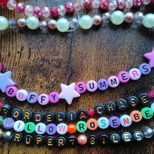 May include: A collection of beaded necklaces featuring a variety of colourful beads and star-shaped charms. One necklace spells out "BUFFY SUMMERS" using letter beads. Other necklaces display names like "CORDELIA CHASE ROSENBERG" and "WILLOW GILES".