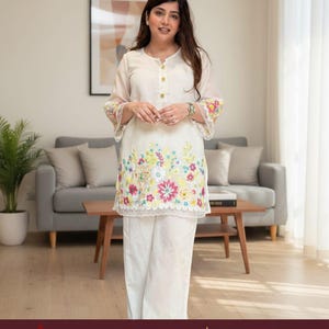 May include: A white long-sleeved tunic featuring a round neckline with three gold buttons and colorful floral embroidery along the hem and cuffs, paired with matching white wide-leg trousers. The garment has a scalloped lace trim at the hem.