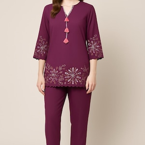 May include: A plum-colored two-piece outfit featuring a tunic top and pants. The top has a V-neck with a tassel necklace, three-quarter sleeves, and embroidered floral details. The pants are straight-legged. The outfit is suitable for casual or semi-formal wear.