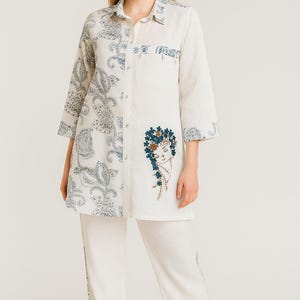 May include: A two-piece white outfit featuring a long-sleeved tunic and matching pants. The tunic has a paisley print on one side and an embroidered face design. The pants have a subtle design along the side. The outfit is worn with black heels.