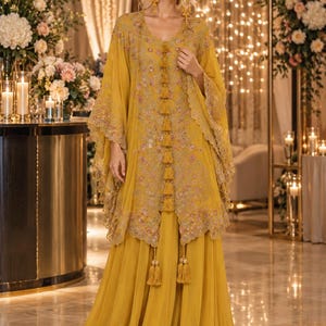 May include: A mustard yellow kaftan-style tunic featuring intricate floral embroidery, scalloped edges, and a vertical row of decorative tassels down the center. The tunic is paired with a matching floor-length, wide-leg skirt in the same shade.