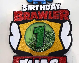 May include: A birthday cake topper featuring a blue and white cartoon character on top. The topper has the words "BIRTHDAY BRAWLER" and "ELIAS" in red glitter. A green number 1 is in the center.