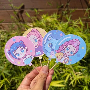 May include: Four colorful, round paper cutouts of anime-style characters on wooden sticks. Each character is depicted inside a balloon shape, with different hair colors and outfits. The cutouts are held against a background of green foliage.