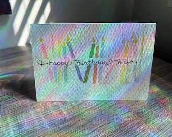 May include: A birthday card with the message "Happy Birthday To You!" written in black script. The card features colorful, painted candles with yellow flames. The card is on a wooden surface with rainbow light reflections.