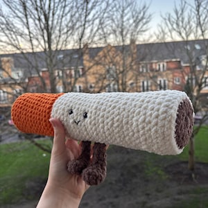 Könnte beinhalten: Handgefertigtes Häkeltier in Zigarettenform mit orangefarbenem Filter, weißem Körper und braunem Ende. Das Spielzeug hat kleine schwarze Augen, ein gesticktes Lächeln und braune Beine.