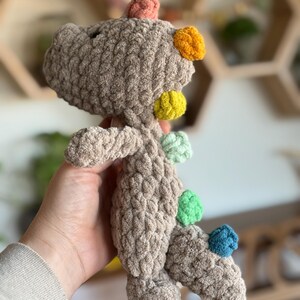 May include: A brown crocheted dinosaur toy with colorful pom-poms on its back. The pom-poms are orange, yellow, green, and blue.