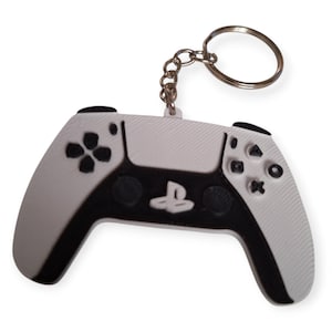 May include: A white and black video game controller keychain. The controller features a black central panel with the PlayStation logo and black buttons. A silver key ring and chain are attached to the top of the controller.