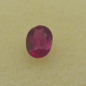 以下が含まれることがあります： ファセット加工された楕円形のルビーの宝石。宝石は深く豊かな赤色で、光を捉える複数のファセットがあります。ルビーは薄い黄色の背景に配置されています。