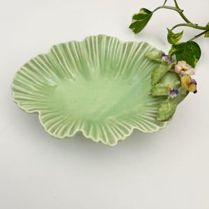 Op de afbeelding: Een lichtgroene keramische schaal met een geschulpte rand, in de vorm van een blad. De schaal is versierd met kleine bloemendetails in paars, geel en crème. Een takje groen-wit gevlekt blad voegt een natuurlijk tintje toe.