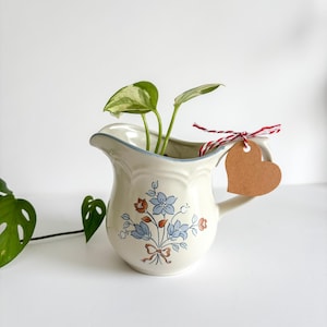 Puede incluir: Jarra de cerámica color crema con un diseño floral en azul y naranja, y un borde azul. Dentro hay una pequeña planta con hojas verdes y blancas. Una etiqueta en forma de corazón está atada al asa con hilo rojo y blanco.
