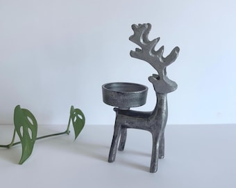 May include: A silver-colored metal reindeer candle holder. The deer has large antlers and a circular holder for a candle. Green leafy vines are in the background. The candle holder is a decorative item.