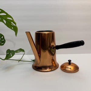 May include: A copper-colored coffee pot with a black handle and a matching lid. The pot has a spout and a rounded base. The handle is made of a dark material. The lid is round with a small knob. The pot is sitting on a white surface.