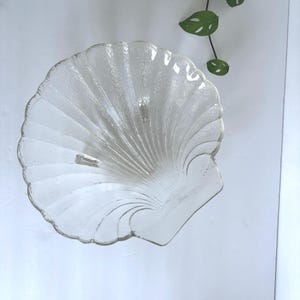 May include: A clear, textured glass seashell-shaped dish. The dish has a scalloped edge and a textured surface, with radial lines emanating from the center. The dish is designed to resemble a seashell.
