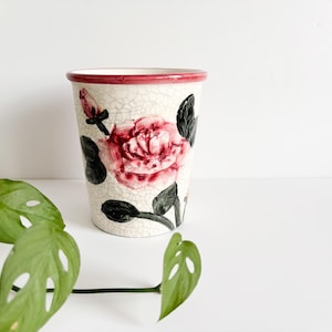 Könnte beinhalten: Eine Keramikvase mit Craquelé-Finish, verziert mit einer großen roten Rose und schwarzen Blättern. Der Rand ist rot. Ein dekoratives Element für die Inneneinrichtung, ideal für Blumen.